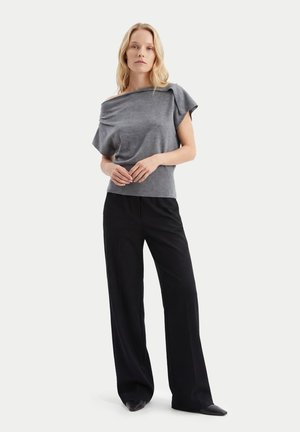 Frau trägt ein graues schulterfreies Kurzarmtop mit schwarzen weiten Hosen und schwarzen spitzen Schuhen, steht mit verschränkten Händen.