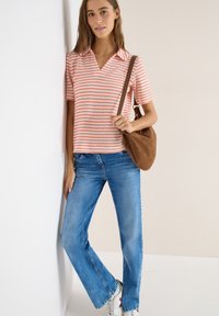 Femme appuyée contre un mur portant un t-shirt rayé à manches courtes, un jean bleu, des baskets blanches et un sac à bandoulière marron.