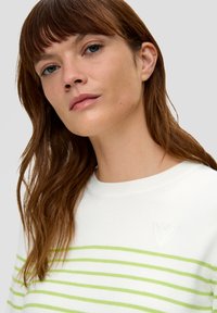 Weißer Baumwollpullover mit grünen horizontalen Streifen, einem Bootsausschnitt und einem kleinen, herzförmigen gestickten Detail auf der Brust.