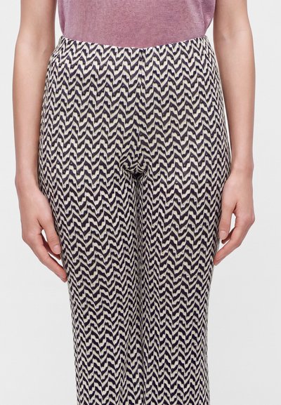Personne portant un pantalon taille haute avec un motif en zigzag noir et blanc et un haut mauve rentré, les mains détendues sur les côtés.