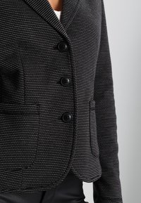 Černobílý pruhovaný blazer s šálovým límcem, třemi černými knoflíky a dvěma bočními kapsami, vyrobený z texturované látky.