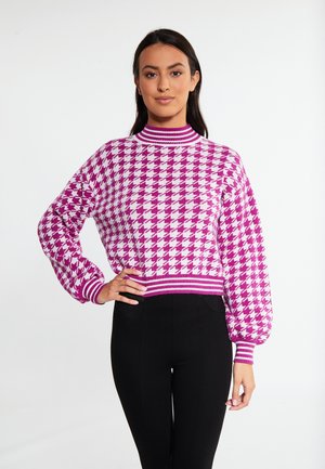 Donna che indossa un maglione rosa e bianco con motivo pied de poule e maniche a palloncino, abbinato a pantaloni neri, in piedi davanti a uno sfondo bianco.
