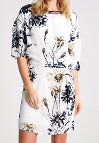 Blommig klänning i vitt med blå och guldiga accenter. Korta ärmar, båtformad halsringning, A-linjeform, med stora prästkragormönster.