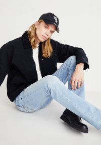 Svart denimjacka med dragkedja, vit t-shirt, ljusblå jeans, svart keps med logotyp och svarta ankelboots. Modell sittande mot en neutral bakgrund.