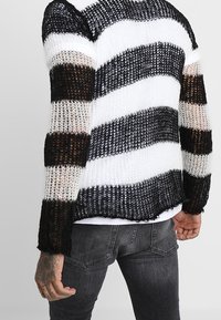 Maglione a righe nero, bianco e marrone, lavorato a maglia con un tessuto allentato, scollatura rotonda e orlo grezzo, indossato sopra una camicia bianca.