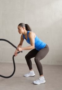 Pessoa com uma t-shirt sem mangas azul e leggings castanhas realiza um agachamento enquanto segura uma corda preta numa academia com um fundo cinza claro.