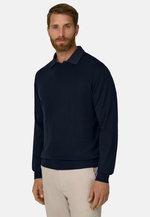 Homme barbu portant un pull bleu marine foncé par-dessus une chemise colletée et un pantalon beige clair, debout devant un fond uni et clair.