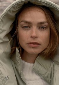 Giacca imbottita grigia chiara con cappuccio, dotata di colletto a coste e zip, indossata sopra un maglione lavorato a maglia bianco. Trucco naturale e capelli disordinati.