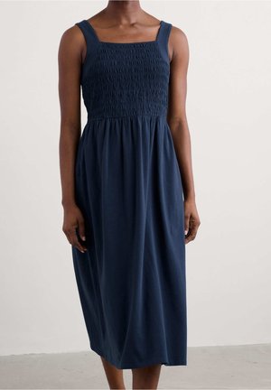 Femme portant une robe midi bleu marine sans manches avec un corsage froncé et un décolleté carré, debout devant un fond clair uni.