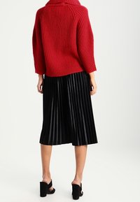 Rode gebreide trui met een hoge kraag en een geribbelde textuur, in combinatie met een zwarte plissé midi rok en zwarte open teen haksandalen.