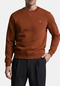 Sweat-shirt marron en tissu lisse, avec un col rond, des manches longues et un petit logo sur la poitrine.