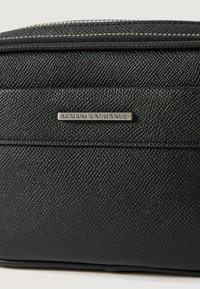 Czarna torba syntetyczna o teksturowanej powierzchni z metalową plakietką "Armani Exchange" z przodu oraz zapięciem na zamek błyskawiczny.