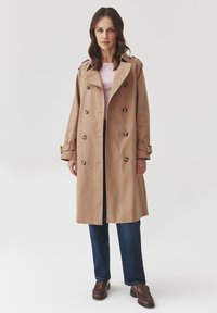 Beiger Trenchcoat mit doppelt bruster Gestaltung, großen Knöpfen, breiten Revers und verstellbaren Manschetten. Getragen über einem hellrosa Oberteil und blauen Jeans.