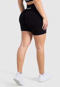 Zwarte sportshorts met een hoge tailleband gemarkeerd met "SMILODON" en een fitted ontwerp, gecombineerd met witte sneakers. Glad, rekbaar materiaal.