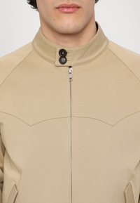 Béžová bunda s vysokým límcem, předním zipem a dvěma knoflíky na krku. Hladká textura s minimalistickým designem a bez viditelných vzorů.