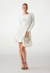 Robe blanche en tricot avec manches longues et jupe plissée. Le modèle tient un petit sac à main beige. Portée avec des talons marron à lanières.