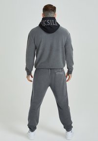 Grijze fleece tracksuit met een zwarte capuchon, met een logo op de achterkant. De pasvorm is relaxed en de textuur is zacht en glad.