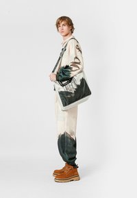 Bolsa tote gris y verde con estampado escénico, que cuenta con una correa resistente y tela texturizada. El modelo lleva una camiseta gráfica y pantalones jogger beige.