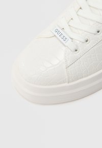Baskets blanches avec un motif crocodile texturé, bout rond et lacets blancs ; marquées du logo "GUESS" sur le côté, semelle en caoutchouc plate.