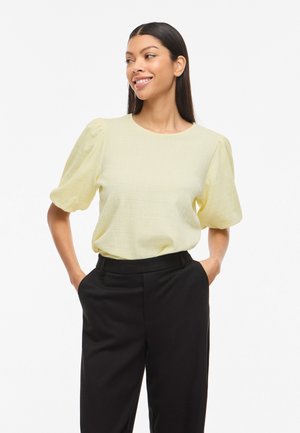 Kvinde smiler og kigger til siden iført en lysegul bluse med pufærmer og sorte bukser med høj talje med hænderne i lommerne.