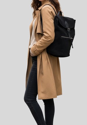 Zaino nero con tessuto testurizzato, dettagli in pelle e chiusura a pattina. Indossato con un cappotto beige, maglione crema e jeans neri.