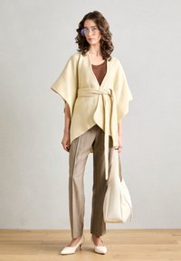 Beige omslagnamn med mjukt material och knyt i midjan, bära över en brun topp och matchande grårandiga byxor samt krämfärgade klackar.