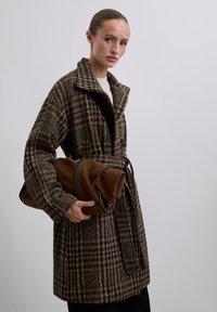 Abrigo marrón a cuadros con un amplio collar, cintura ceñida y dos botones; sosteniendo un bolso de ante marrón con una textura suave.