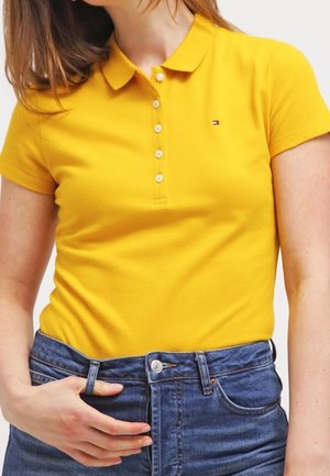 Polo shirt jaune avec un col, doté d'une patte de boutonnage à trois boutons et d'un petit logo. Associé à un jean en denim bleu.