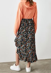 Dlouhá květinová sukně s černým pozadím, která má barevné květinové vzory a volánkový lem, spárovaná s korálovým mikinou a bílými teniskami.