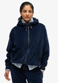 Sudadera azul marino de felpa con cremallera, capucha, bolsillos frontales y puños acanalados. Lleva encima una camiseta azul clara y pantalones con cintura ajustable.