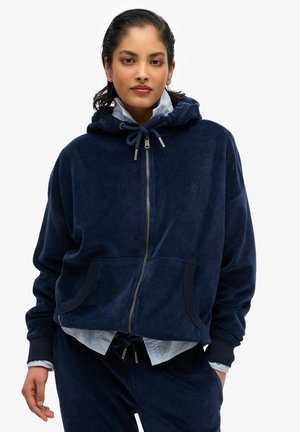 Veste à capuche zippée en velours bleu marine avec une capuche, des poches avant et des poignets côtelés. Superposée à une chemise bleu clair avec un pantalon à taille à cordon.
