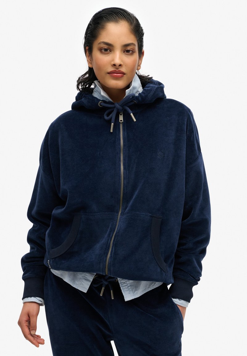 Sudadera azul marino de felpa con cremallera, capucha, bolsillos frontales y puños acanalados. Lleva encima una camiseta azul clara y pantalones con cintura ajustable.
