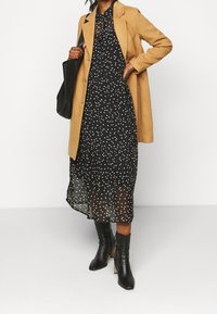 En kamelfärgad kappa över en svart klänning med ett subtilt vitt mönster. Blanka svarta ankelskor och en svart toteväska fulländar looken.