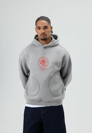 Grijze capuchontrui met een rond rood logo met een roos. Het heeft grote zakken aan de voorkant en een losse pasvorm. Gedragen over een wit shirt.