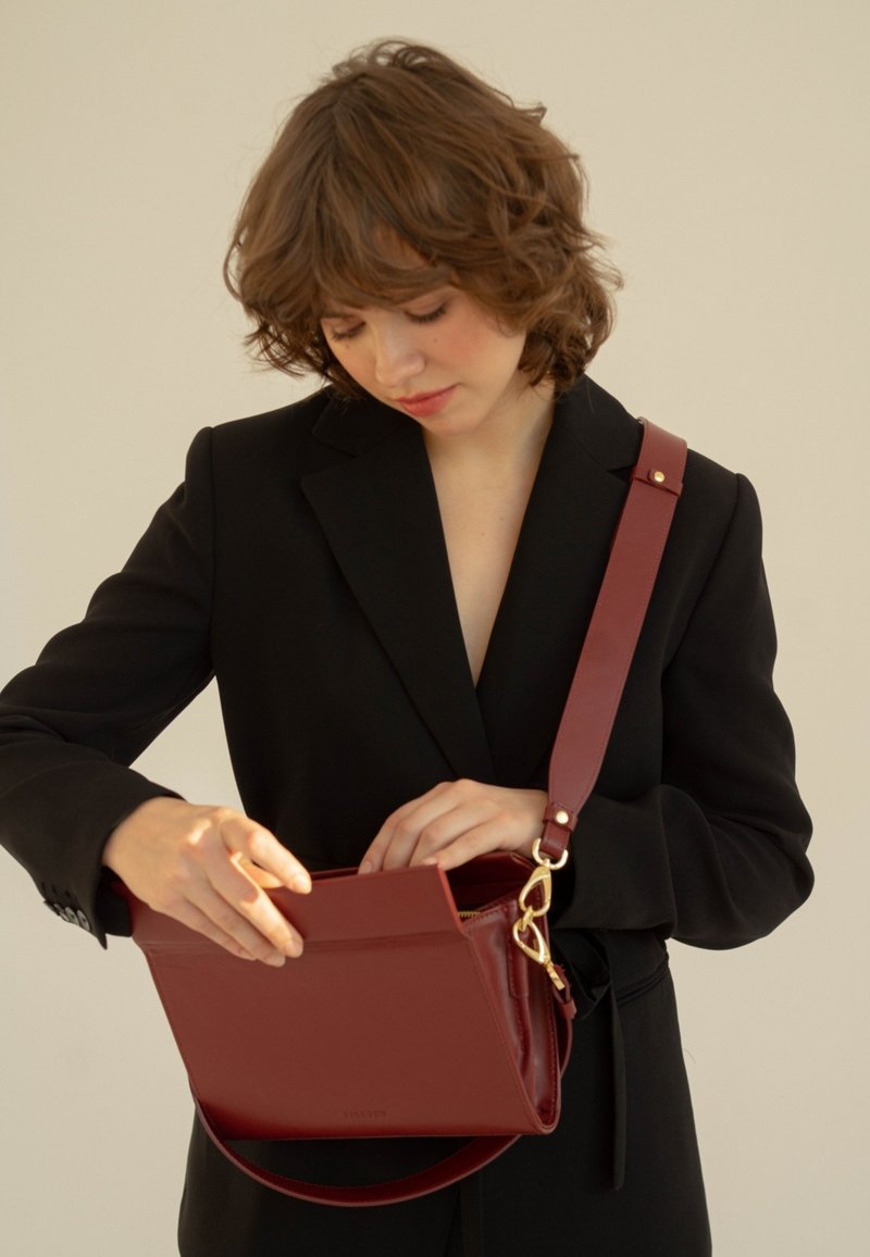 Strukturerad röd läderhandväska med axelrem, med ett topplås i dragkedja och en guldtonad detalj. Slät textur, rektangulär form.