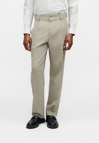 Άνδρας με μπεζ παντελόνια ίσιας γραμμής, λευκό πουκάμισο με κουμπιά και μαύρα loafers, στέκεται μπροστά σε απλό φωτεινό φόντο.