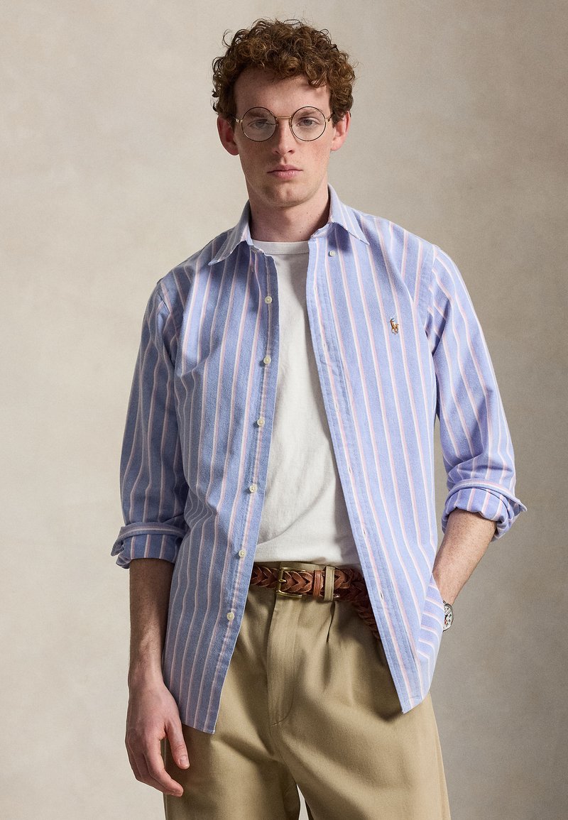 Ung mann med krøllete hår og runde briller iført en blåstripet skjorte med knapper over en hvit t-skjorte, beige bukser og et brunt flettet belte.