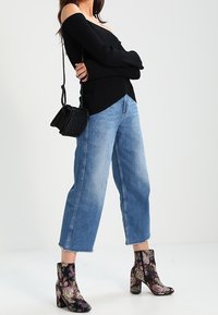 Mujer con un suéter negro sin hombros, jeans azules recortados, botas tobilleras con estampado floral y un bolso cruzado negro con detalle de cadena.