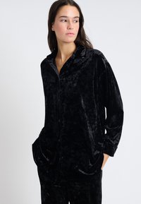 Camisa de pijama de terciopelo negro con cuello, botones en la parte delantera, mangas largas y bolsillos laterales. Textura suave con un acabado ligeramente brillante.