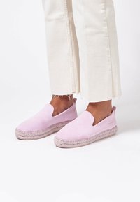 Scarpe slip-on in suede rosa con punta arrotondata, caratterizzate da una suola in juta intrecciata e dettagli laterali a taglio. Abbinate a pantaloni chiari con orlo sfrangiato.