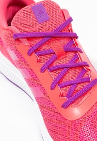 Chaussure de sport rose vif avec une tige en mesh texturé, lacets violets et accents réfléchissants. Présente un logo audacieux sur la languette et une semelle lisse.