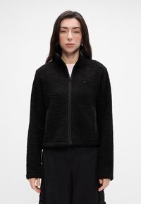 Svart fleecejakke med glidelås foran, høy krage og lange ermer. Mykt, teksturert stoff med logodetalj på brystet.