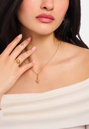 Goldkette mit Anhänger, der einen Edelstein zeigt, getragen zusammen mit einem Goldring. Glatte, helle Haut und natürlicher Nagellack an den Händen. Weißes schulterfreies Oberteil.