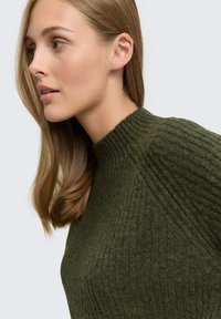Olivgrüner gerippter Strickpullover mit hohem Kragen. Die Textur ist weich und dick, mit vertikalen Streifen und einer figurschmeichelnden Silhouette.