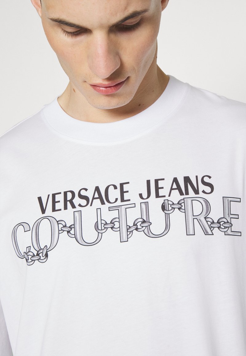 Joven mirando hacia abajo, vistiendo una camiseta blanca con el texto "VERSACE JEANS COUTURE" estilizado con eslabones de cadena en el pecho.