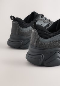 Baskets noires épaisses avec un mélange de dessus en daim texturé et en maille. Semelle extérieure en caoutchouc épaisse avec surpiqûres grises et languette à l'arrière.