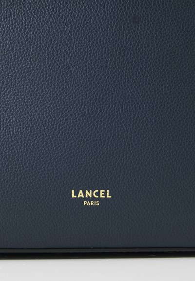 Тъмносиня текстурирана кожа с перлена повърхност; разполага с златно лого "LANCEL PARIS" в долната част.