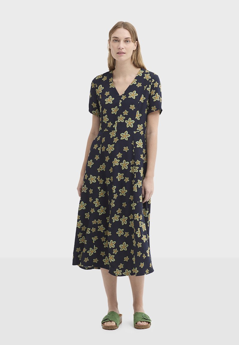Robe bleu marine à motif floral, manches courtes et décolleté en V, ornée de fleurs vertes et jaunes. Associée à des sandales vertes.