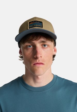 Lichtbruine honkbalpet met een blauwe klep, voorzien van een rechthoekige patch met een berglandschap en de tekst "BILLABONG". Draagt een blauw T-shirt.