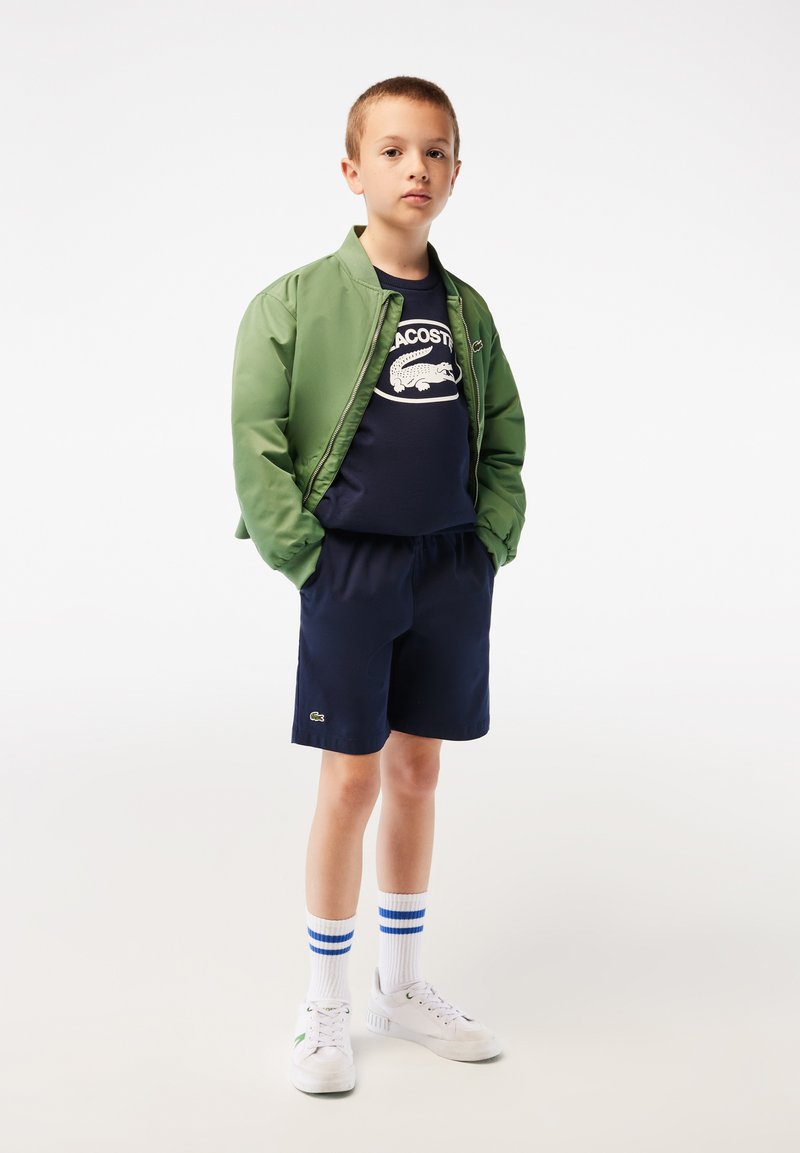Giacca bomber verde, T-shirt blu navy con logo bianco, pantaloni corti blu navy, calze bianche con strisce blu e sneakers bianche con dettagli verdi.